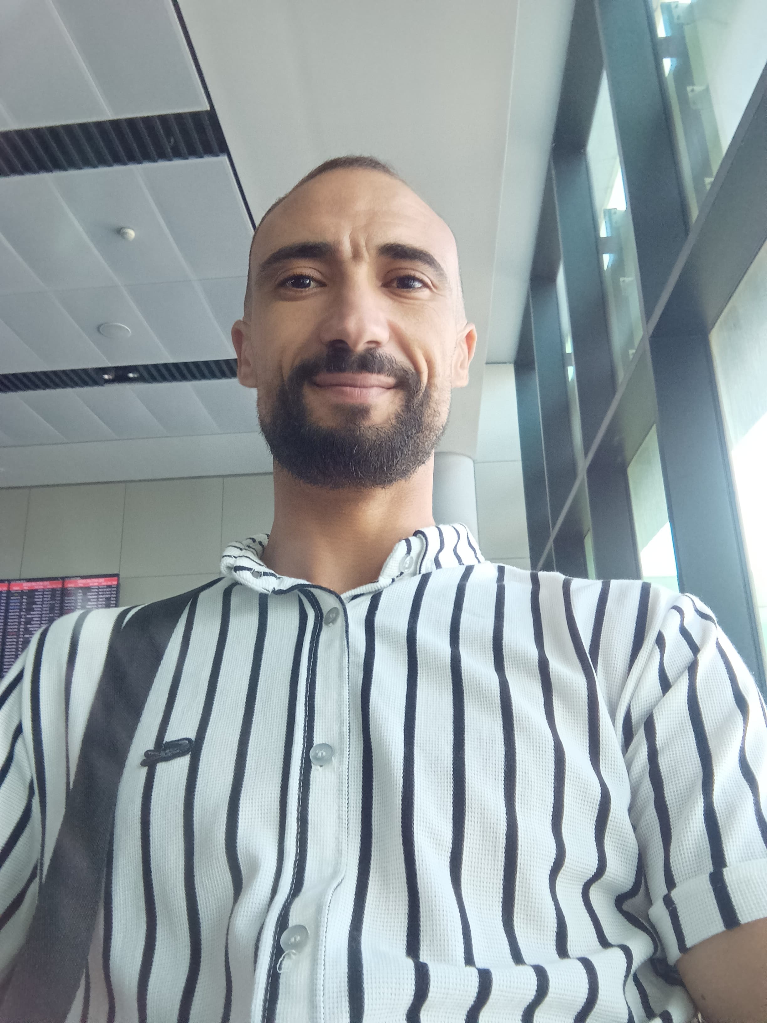 Smiling bearded man wearing a black and white striped shirt in a bright airport terminal.